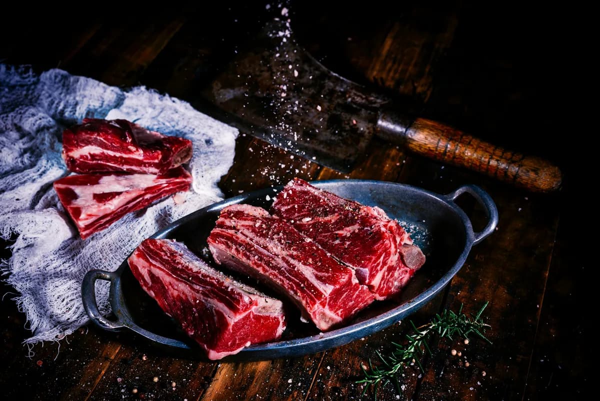 Raw bovine short ribs on a butcher's cutting surface, showing the cortical bone shell and the trabecular interior that make them a realistic natural-bone model for dental implant osteotomy bench training.